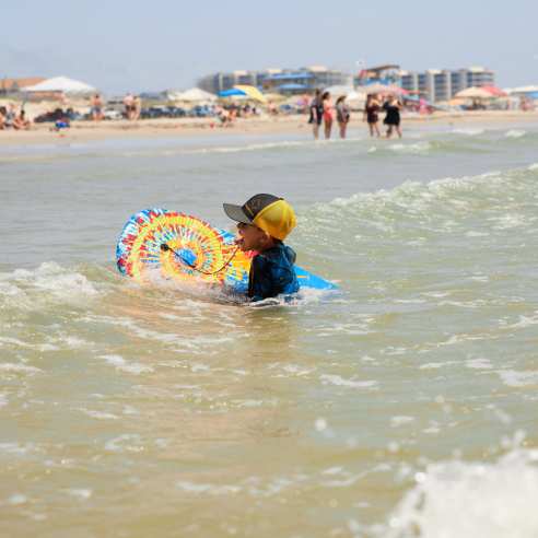 A child in a ball cap swims with a tie-dye boogie board near the beach