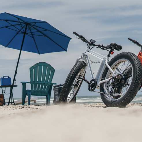 Two bikes, one orange and one white, sit on the beach next to a beach chair setup.