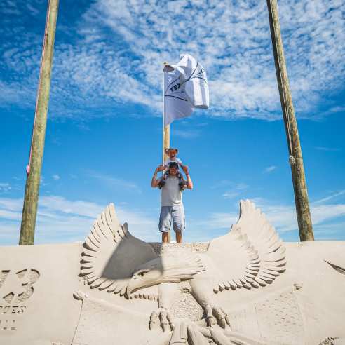 A man carries his toddler daughter on his shoulders and they pose atop a large sand mountain carved with an eagle