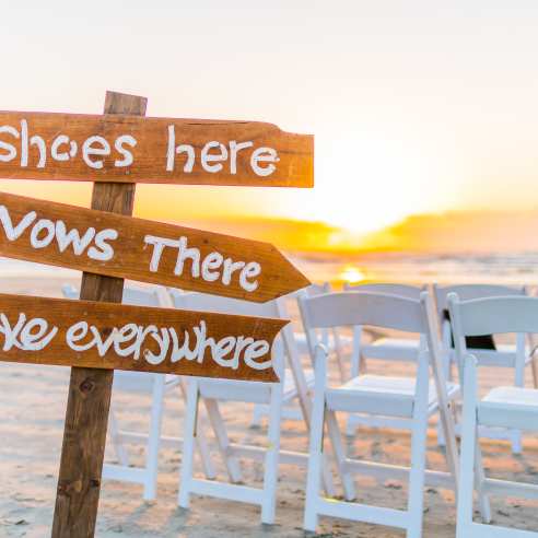 A brown sign is stuck in the sand. It reads "Shoes here," "Vows there," and "Love everywhere."