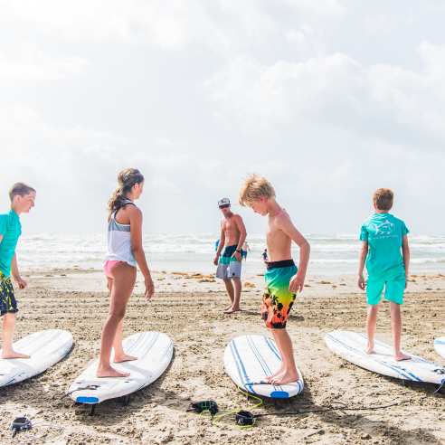 A group of five kids stand on surf boards resting on the sand while an instructor stands in front of them