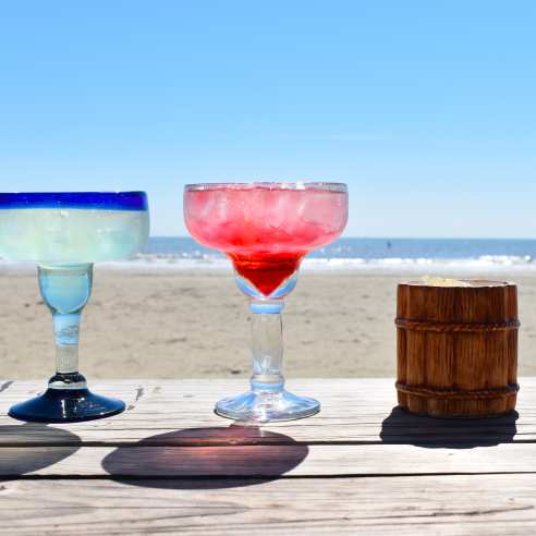 A row of margaritas sitting on a wooden table on the beach.