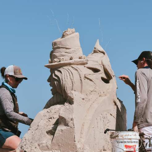 Two people, a man and a woman, stand on either side of a sand sculpture and carve details.