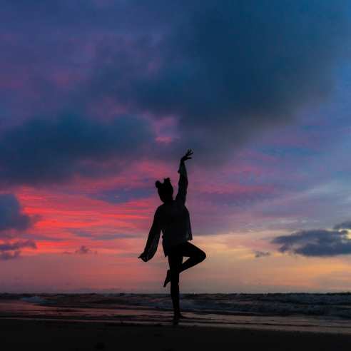 A model is silhouetted in a dancing pose against a dark pink, blue, and purple sunrise on the beach