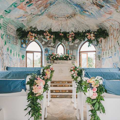 The interior of a chapel painted with murals is decorated with pink, white, and green florals, and white pews are draped in light blue fabric