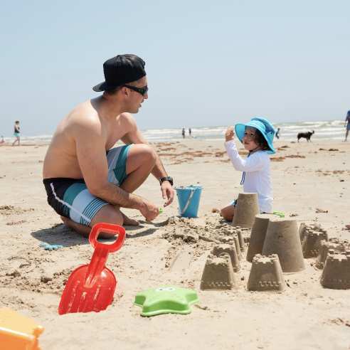 A man in a swimsuit kneels on the sand next to a child wearing sun gear and a hat. They pack sand in a blue bucket and build a sandcastle.