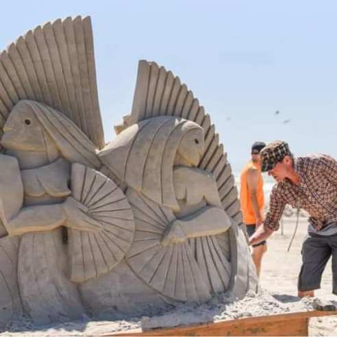 A man in shorts and a plaid shirt bends over a sand sculpture he's working on. The sculpture seem sto depict two figures holding fans, looking away from each other.