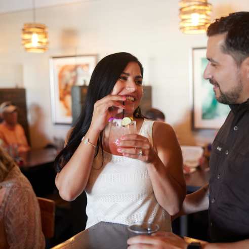A woman with long hair sips on a cocktail and smiles at the man next to her