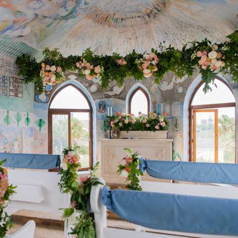 The inside of a small mural-covered chapel is decorated with pink and white flowers and greenery