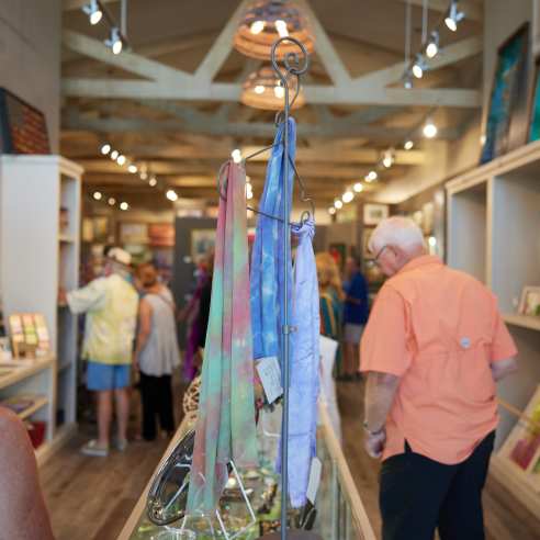 Colorful scarves hang on a rack standing on a shelf in the middle of the photo. Shoppers peruse on either side of the shelf. Paintings hang on the walls on both sides