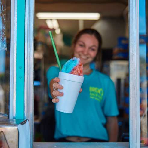 Girl in turquoise shirt holds out a red and blue snow cone in a styrofoam cup outside a window