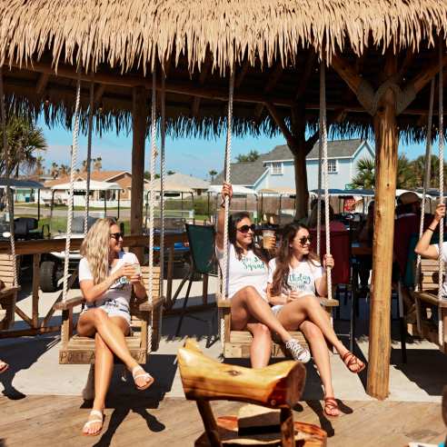 A group of five women wearing matching birthday shirts sit in tiki-style swings