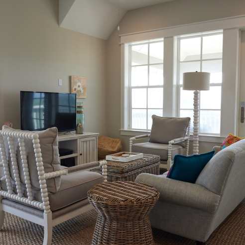 A living room with coastal decor in shades of beige. A tan couch faces a television and two chairs face each other perpendicular to the couch.