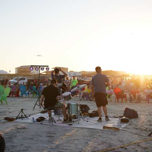 A photo of a three-person band on the beach from behind with a crowd watching from beach chairs