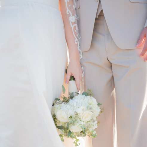 A close-up photo of a couple standing on the beach in wedding attire. The bride holds a bouquet.