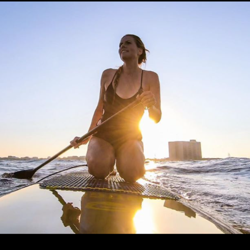Girl kneels on a stand up paddle board in the water with a paddle in hand.