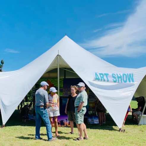 A white tent is pitched in the middle of a grassy field. The tent has blue text reading "Art Show." A group of four adults stand talking in front of the tent.