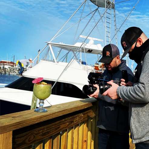 Two men hold large cameras pointed at a tropical margarita on a wooden ledge. A large white yacht is behind them.