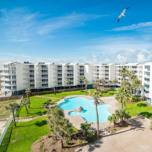 Aerial photo of a beautiful white condominium complex with palm trees, a pool, and green grass.