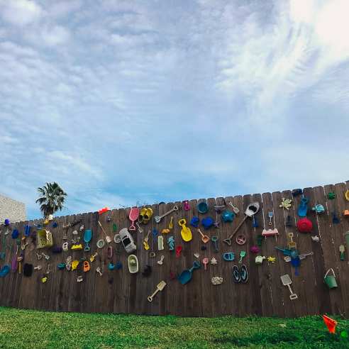 A fence is dotted with colorful discarded beach toys—shovels, cars, buckets, and more. A sky covered in white clouds is above and a house is visible in the left background