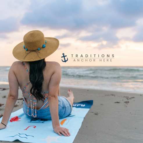 A woman in a bathing suit, shorts, and a beach hat sits on the beach looking out over the water. Next to her is a tagline reading "Traditions Anchor Here" with a dark blue anchor icon.