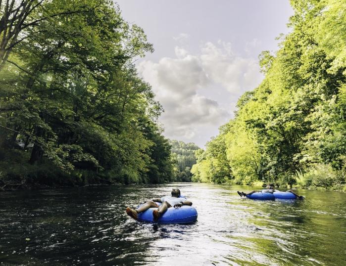 Tour Boone from the river! A calm river with lush green trees on either side has three people in floats, the closest of which has their eyes closed looking up at the sky.