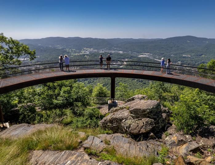 A curved, metal walkway stands on a rocky mountain peak surrounded by green trees, with three couples taking in the views of a mountainous horizon and the Town of Boone below.