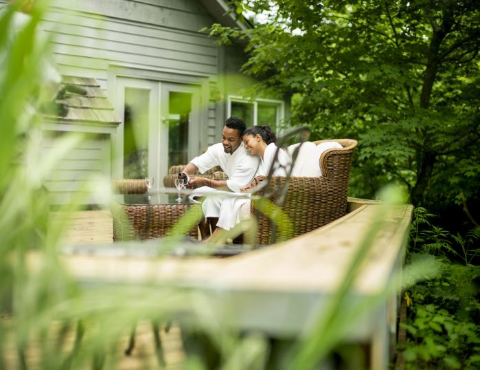 A couple toasts with wine glasses in white robes on a wicker couch outdoors.