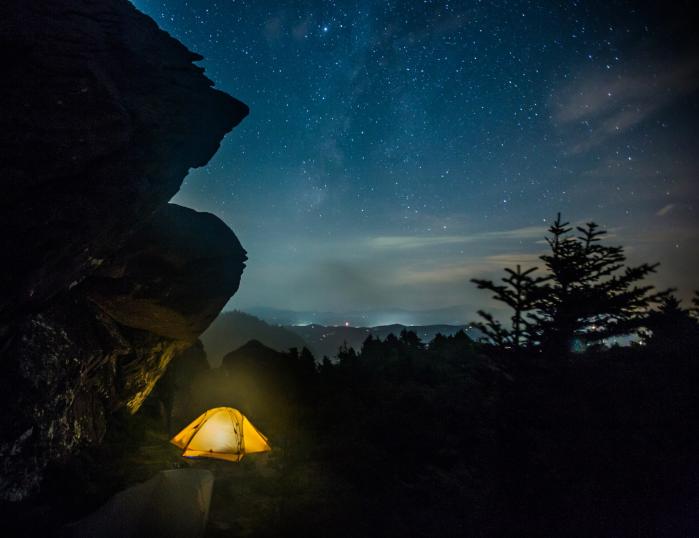 A tent placed below a rocky outcropping emits a yellow glow, with stars appearing in the nighttime sky