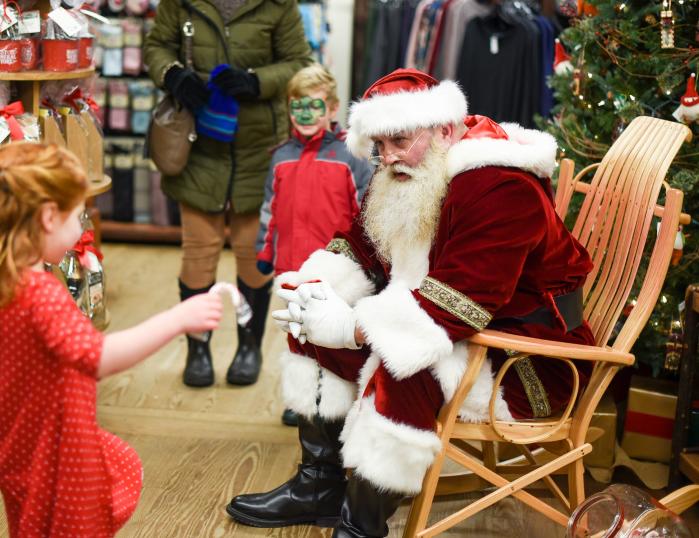 Santa Claus sits in a wooden rocking chair at Mast General Store, as a little girl in a red dress approaches with a candy cane.