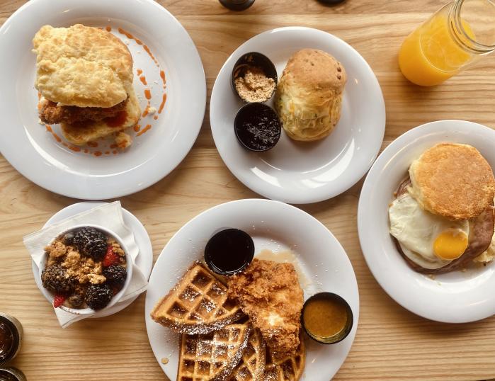 Four white plates and one bowl are piled high with breakfast biscuit sandwiches, chicken and waffles, yogurt, and jams.