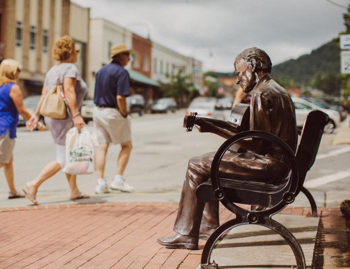 A trio walks with shopping bags past a bronze statue of a man, Doc Watson, playing a guitar on a bench in Downtown Boone.