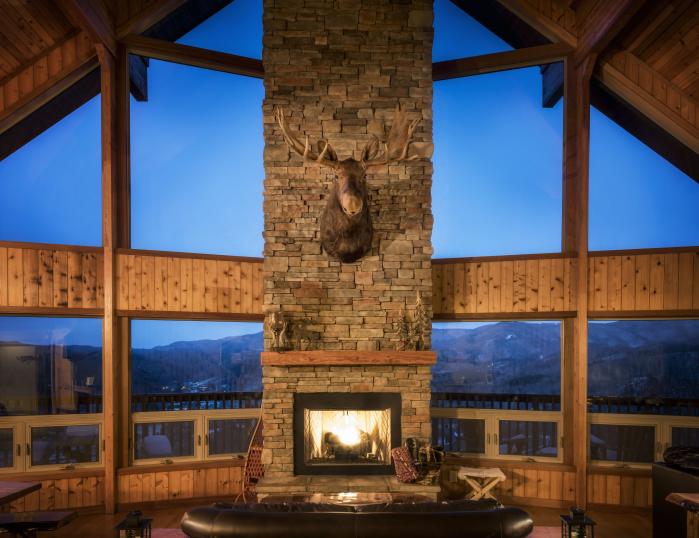 Inside a cabin looking out large windows to a mountain landscape. A fireplace is lit brightly.