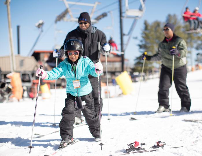 A family, with a small child in a blue jacket and ski poles in both hands in front, makes their way towards the camera.