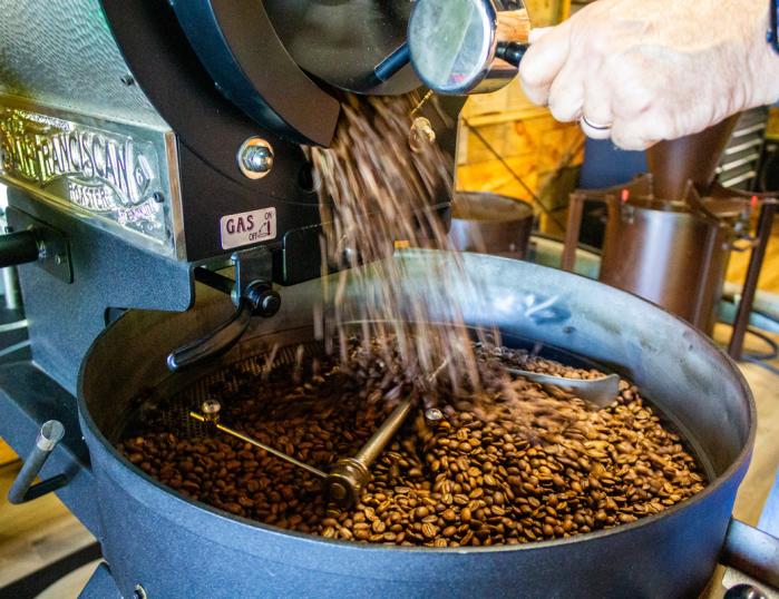 A hand can be seen holding a lever that releases freshly roasted coffee beans, which are pouring from the spout and collecting in the container below.