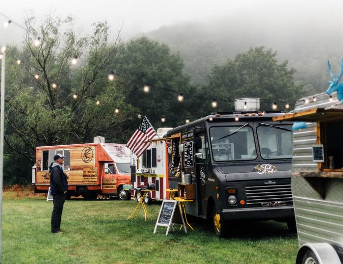 Four food trucks lined up in a grassy field, with a customer reading the menu for 'Betty's Biscuits.'