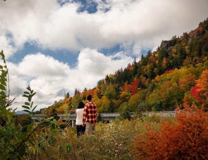 A woman in a white sweater and a man in red flannel view the beautiful fall foliage from the side of the Blue Ridge Parkway.
