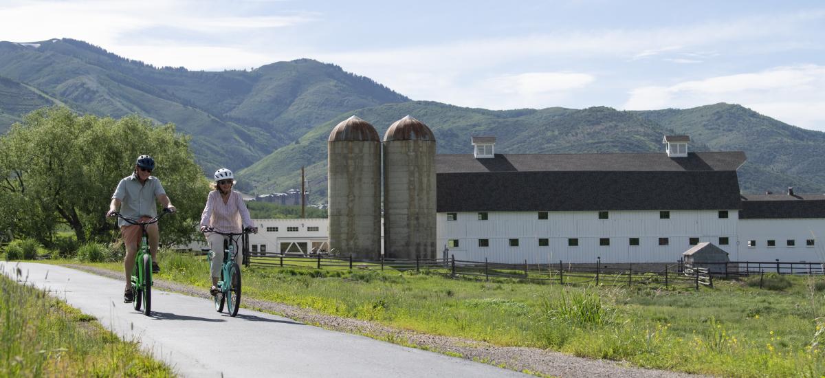 Two people biking a paved trail with the white McPolin Farm in the background.