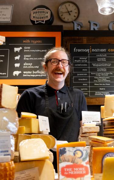 A white man stands behind the counter at Fromagination with a big smile on his face