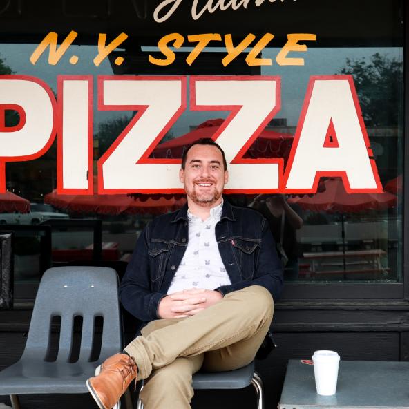 Man sitting on plastic chair in front of painted window with the words "N.Y. Style Pizza".