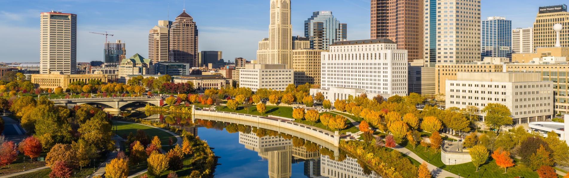 Columbus skyline reflecting in the Scioto River, with red and orange trees lining the river