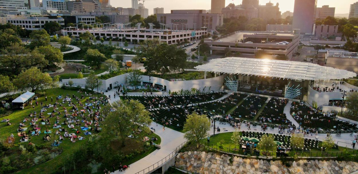 Aerial view of Moody Amphitheater in evening sun. There is a crowd on the open lawn and in seats near the pavilion stage. The State Capitol building is visible behind the amphitheater