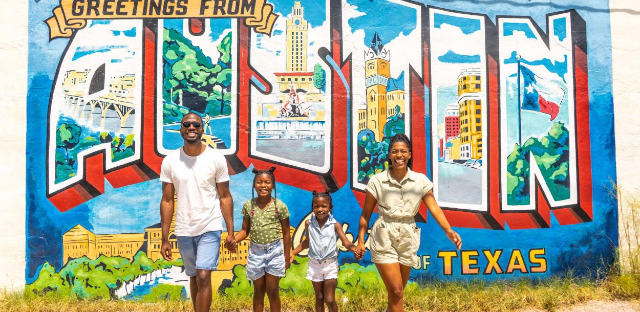 Family of four with two young daughters hold hands and smile at camera in front of postcard mural that reads Greetings from Austin Texas