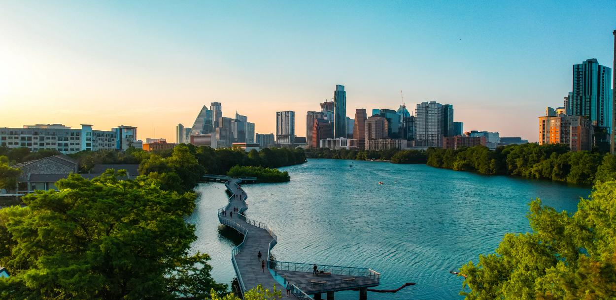 People walking along suspended board walk that follows the shape of the Colorado River, leading to Downtown Austin skyline.