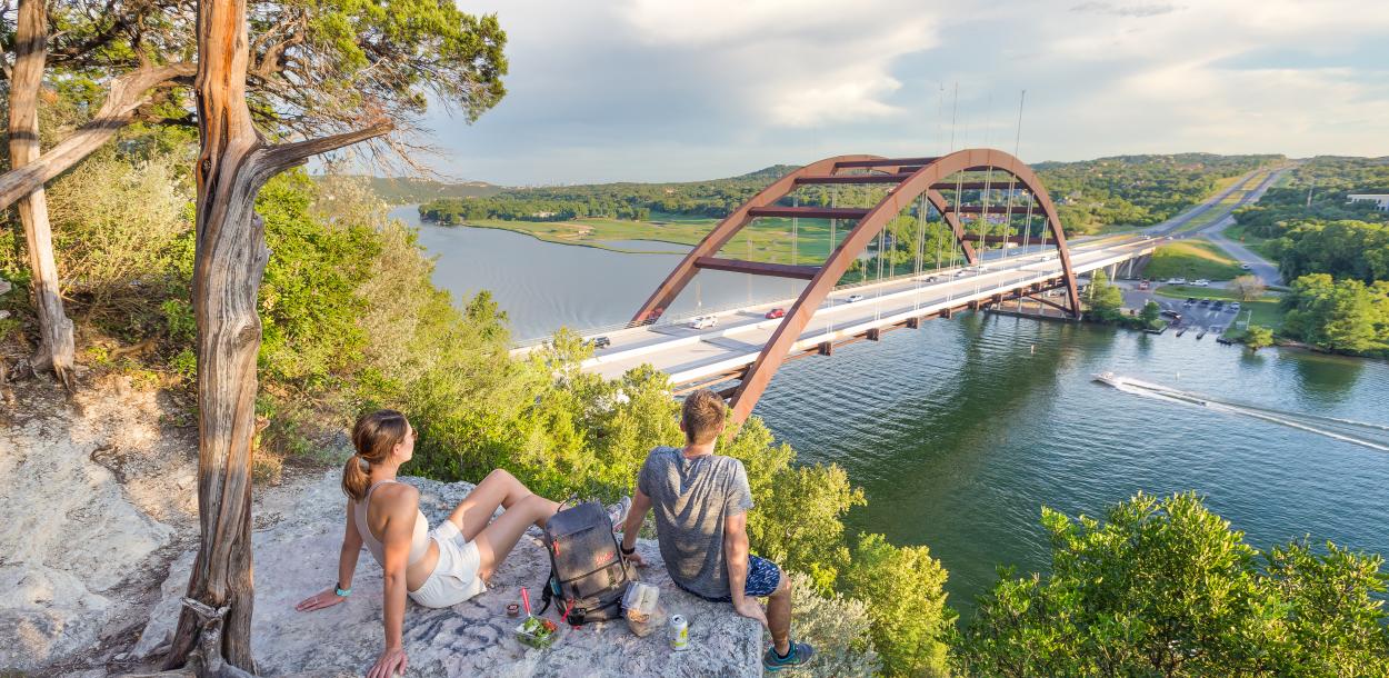 Hikers sitting on a hilltop rock overlooking the Pennybacker Bridge at Lake Austin.