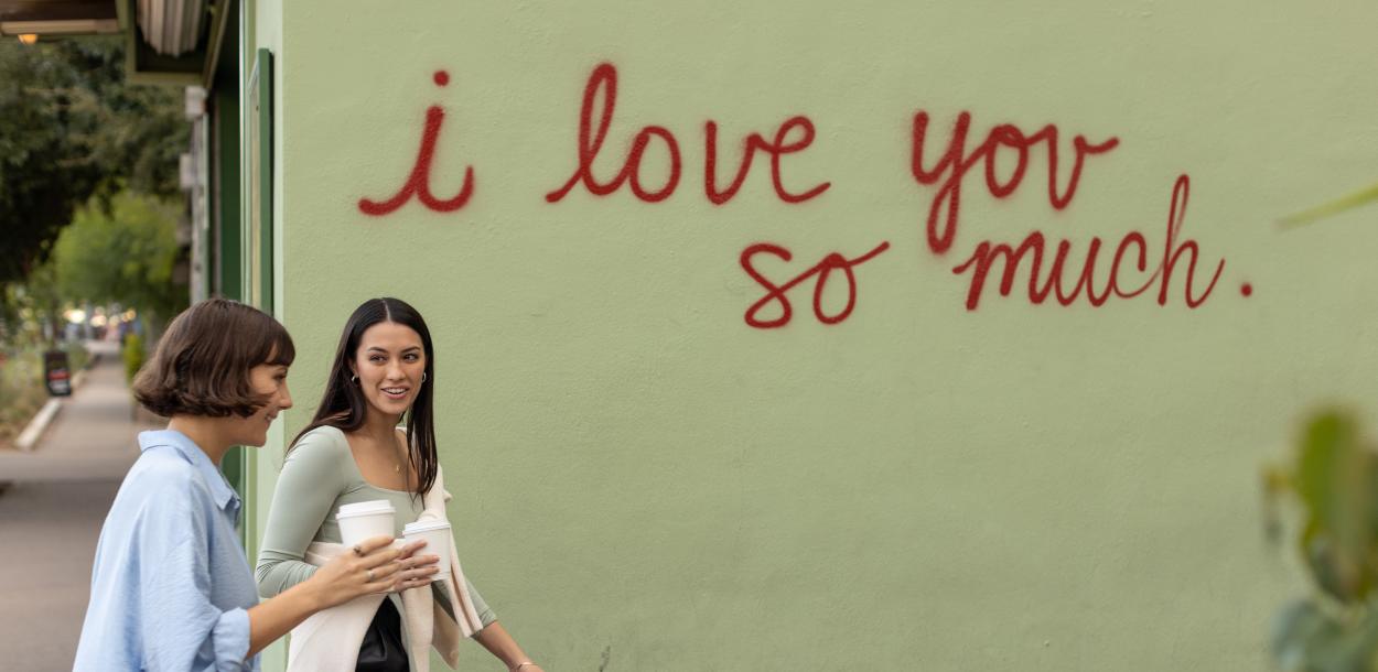 Two women walking in front of the I Love You So Much mural with coffee in hand.