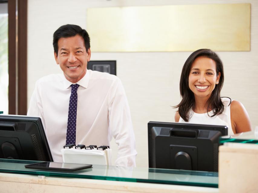 Hotel front desk staff smiling and welcoming guests during check-in at a hotel reception desk.