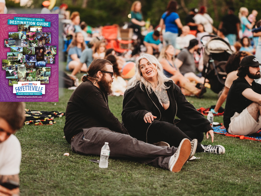 A smiling couple relaxes on a blanket at an outdoor fall festival in Fayetteville, North Carolina, surrounded by people enjoying live music and community activities. A cover image of the Fayetteville Destination Guide appears in the corner, inviting visitors to plan their trip.