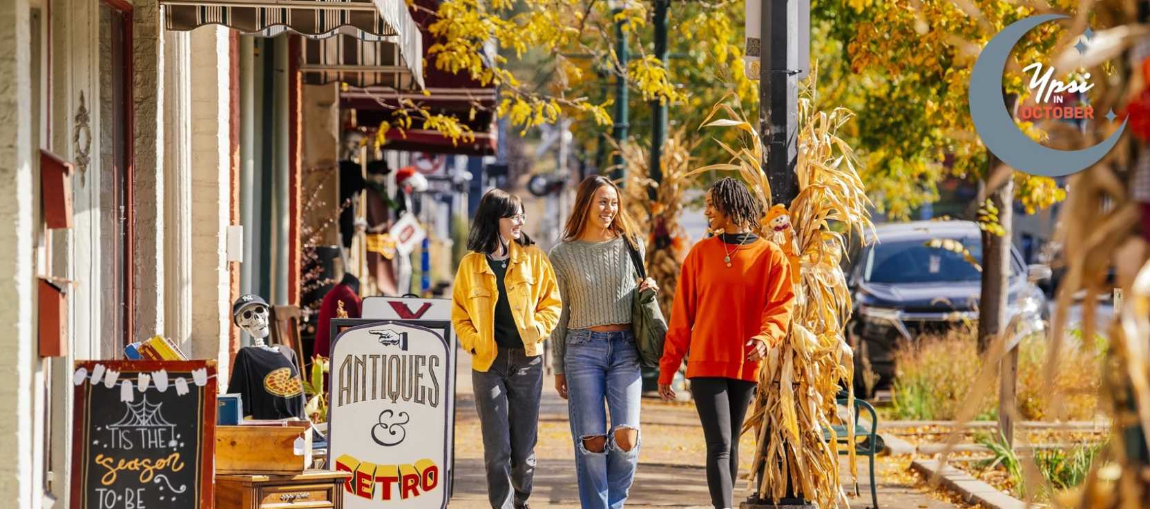 Three women shopping in Depot Town, walking along storefronts on a sidewalk during the fall season. Yellow and orange leaves in the background.