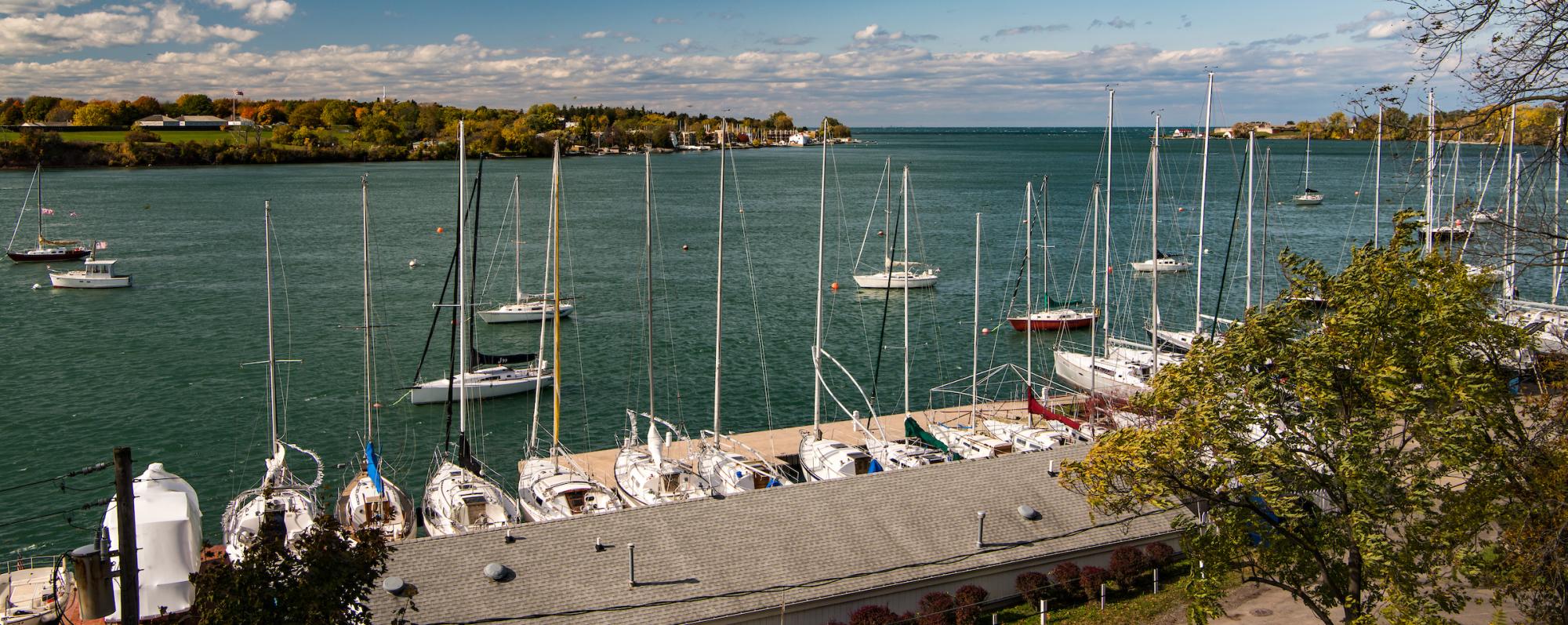 A marina with numerous sailboats docked along the waterfront, overlooking the Niagara River under a partly cloudy sky. Trees and a road are visible in the foreground.